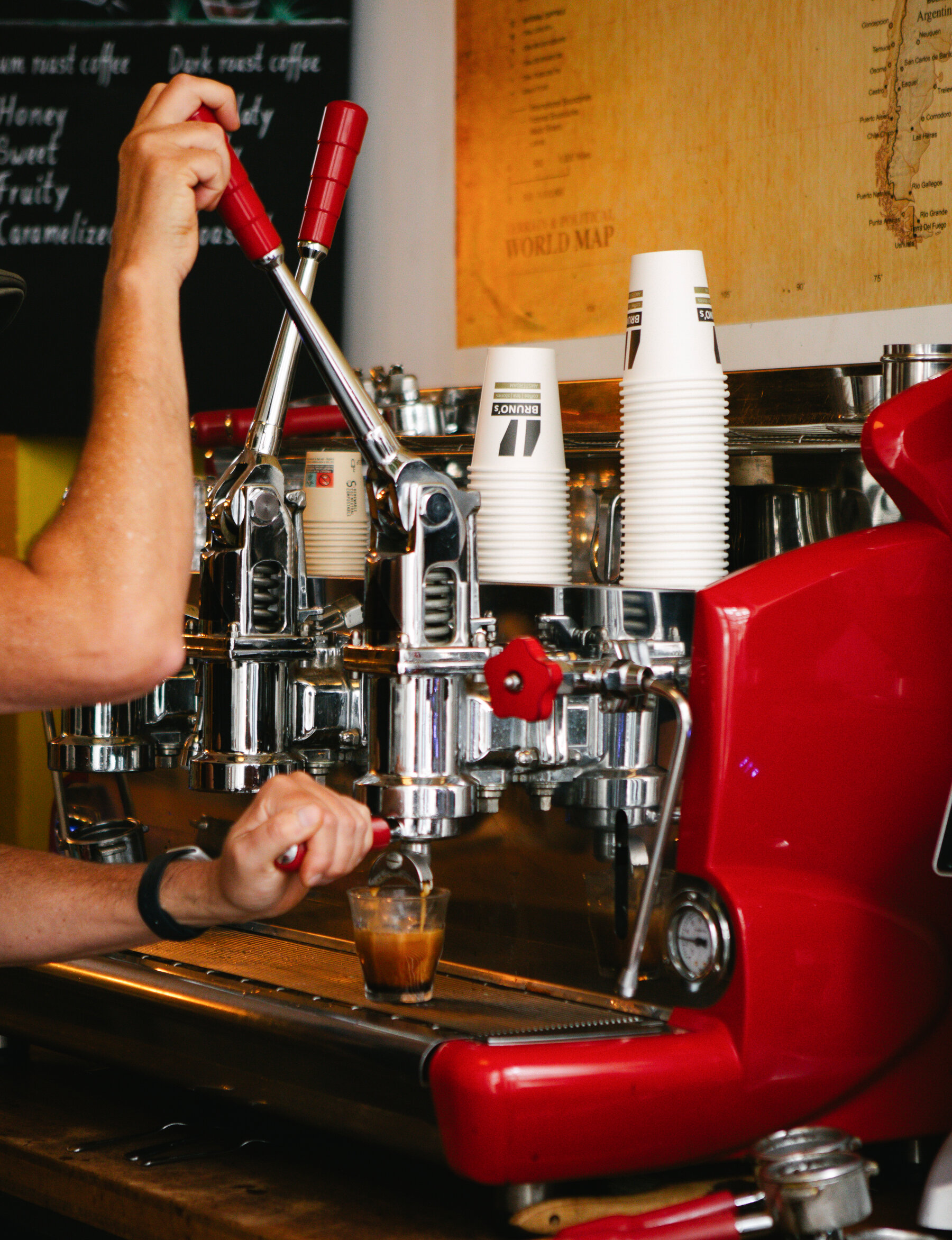 Un aparat de cafea rosu cu un maner mare, argintiu, intr-o cafenea.  O mana intra in cadru si opereaza aparatul, turnand un espresso intr-o ceasca de sticla.