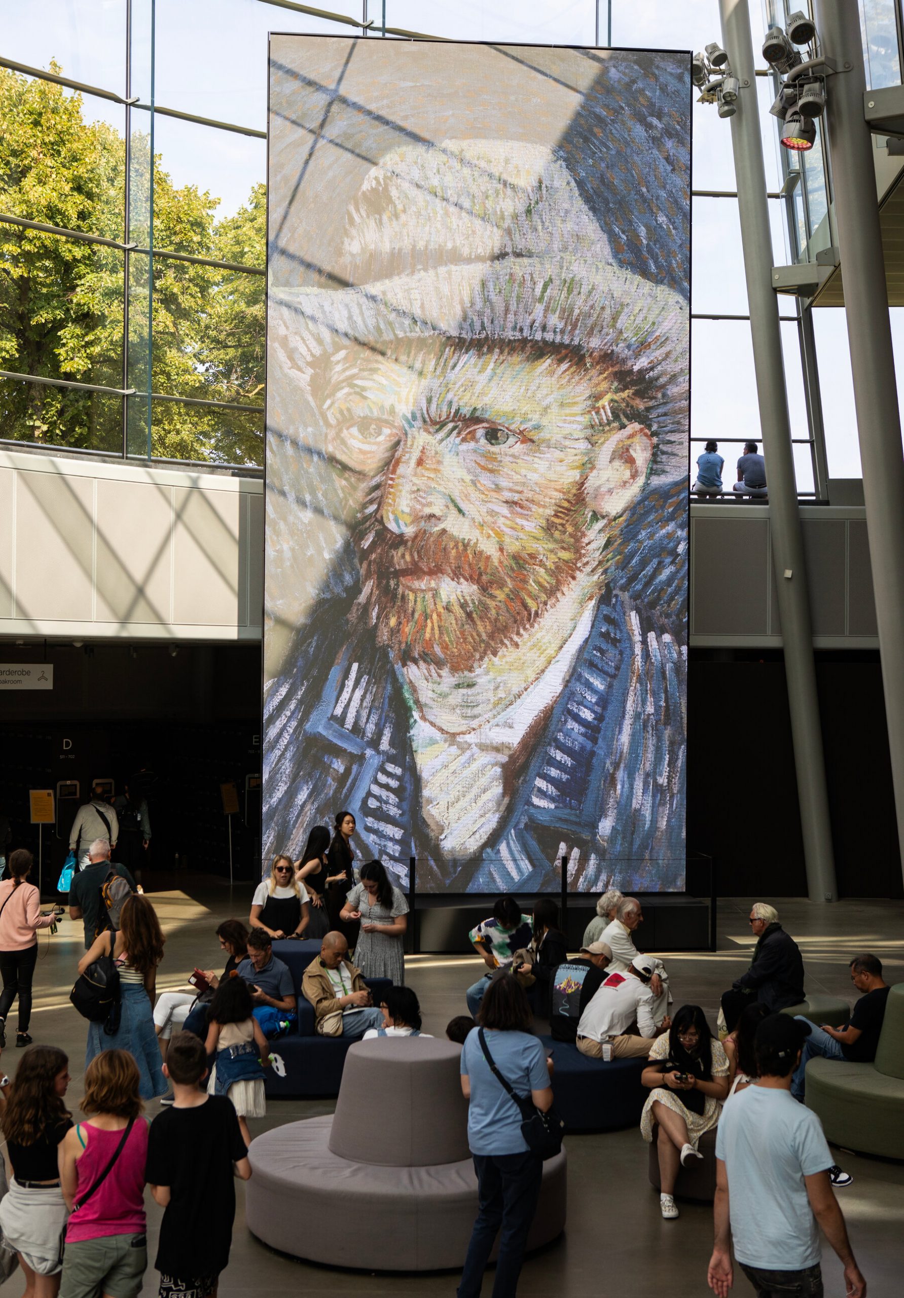 Un foaier al muzeului in care oamenii se plimba si stau pe scaune circulare gri.  O reproducere mare a unui autoportret al lui Vincent van Gogh este afisata in fundal, pe ferestre mari de sticla.