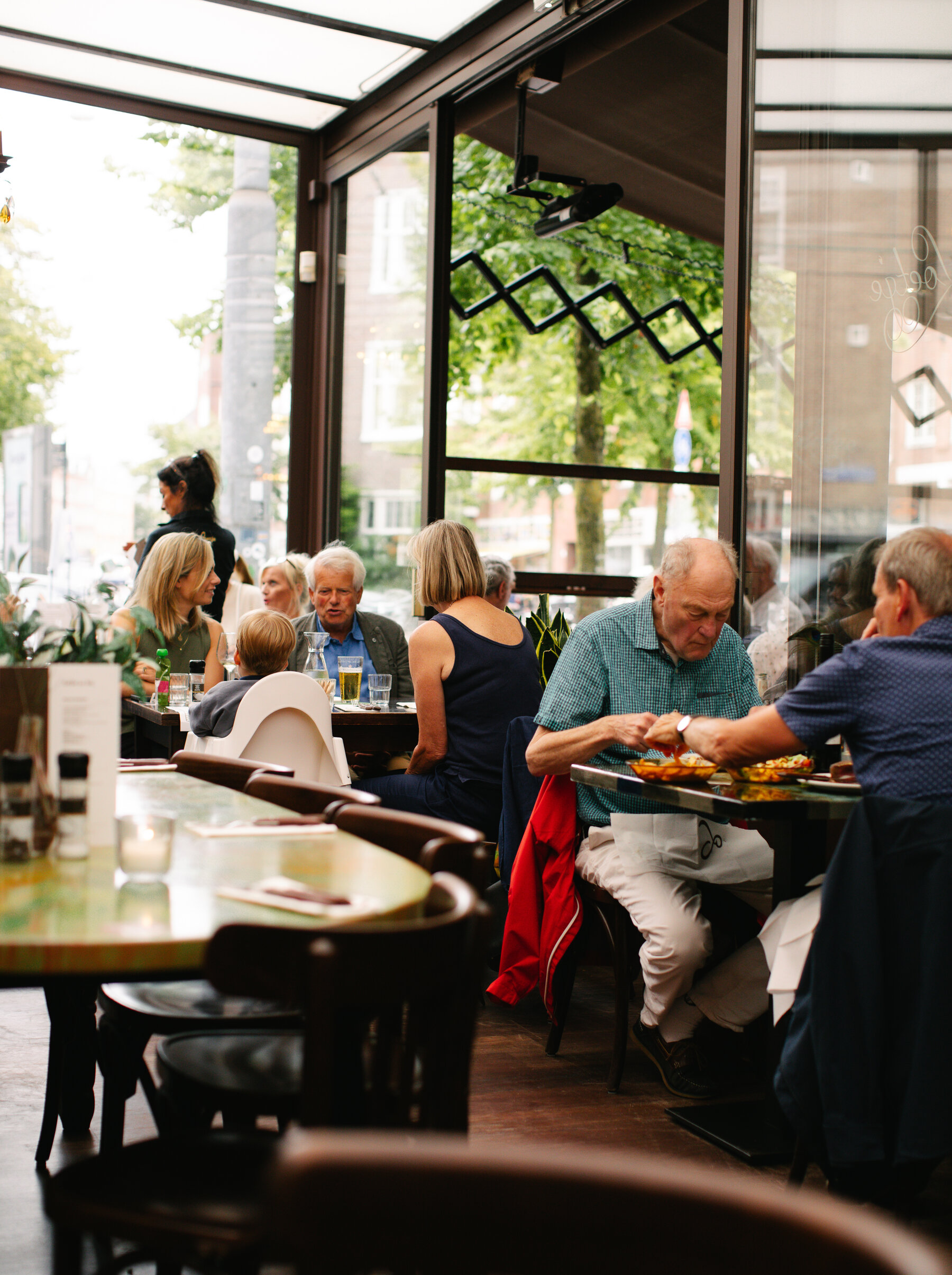 Interiorul unui restaurant cu ferestre mari de sticla care lasa lumina zilei inauntru.  Oamenii stau la mese si scaune de lemn.
